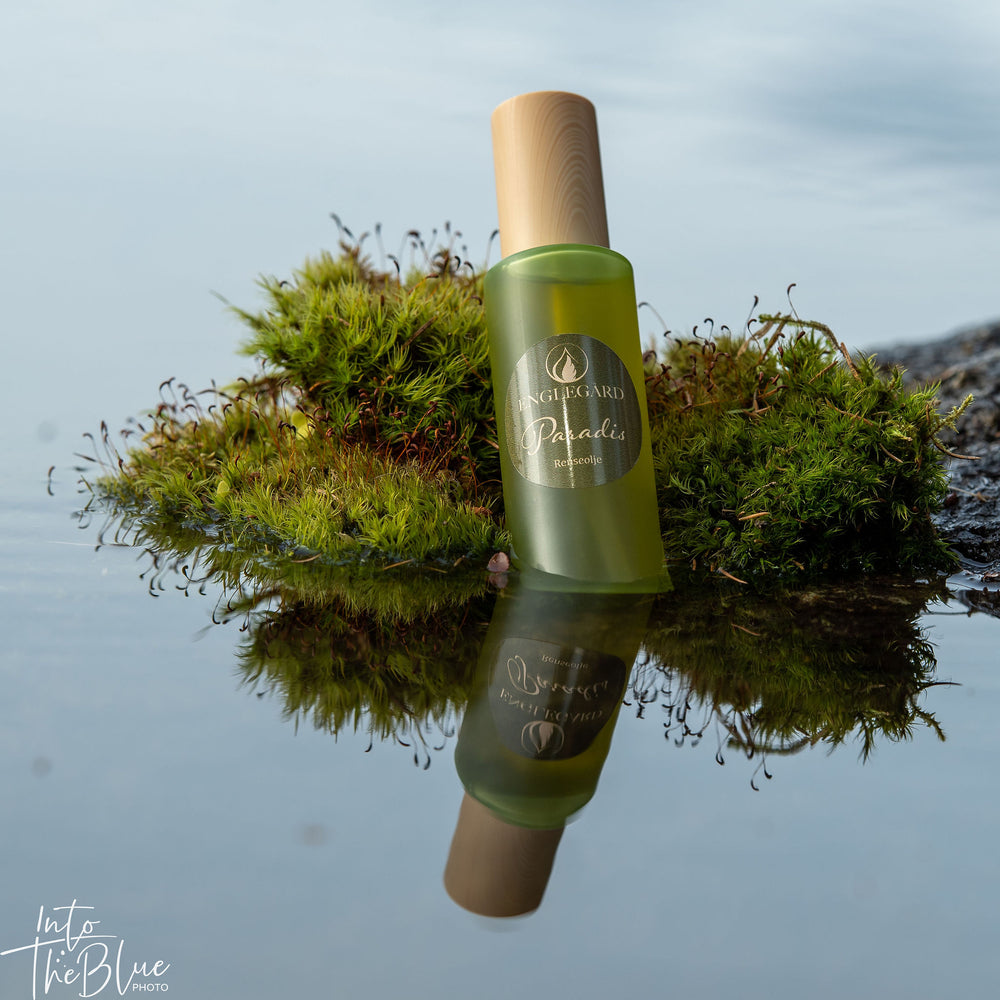 Renseolje, Paradis, fra Englegård i olivenfarget glassflaske med trekork. Fotografert  ved vann med mose som underlag. Flasken speiles i vannet.